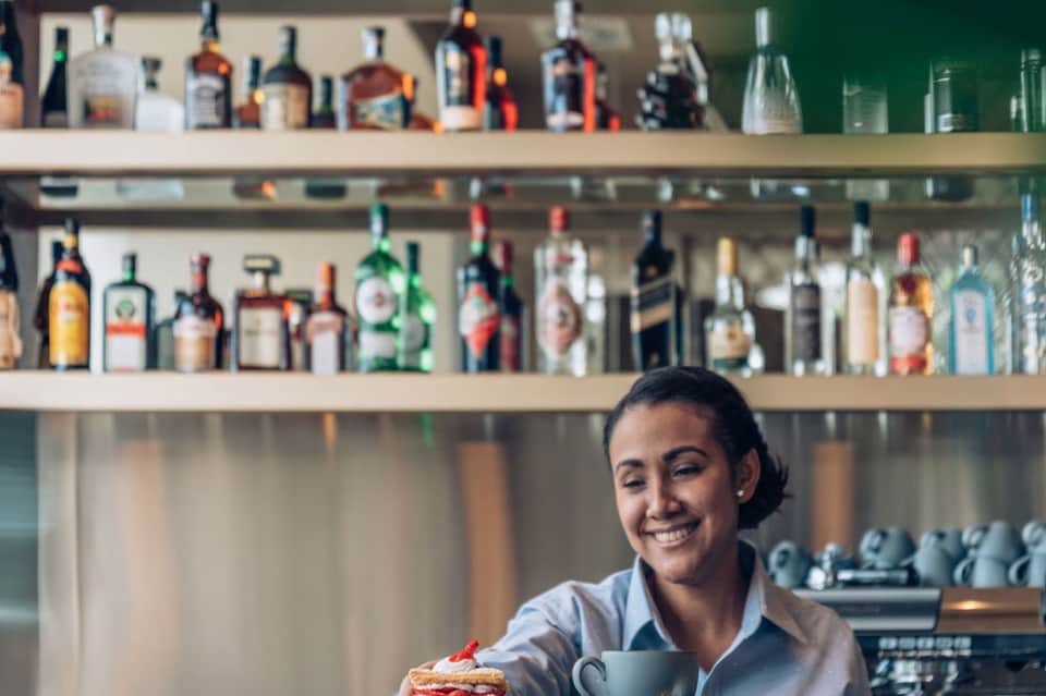 Smiling bartender in white shirt serving dessert at hotel bar with shelves of liquor bottles behind