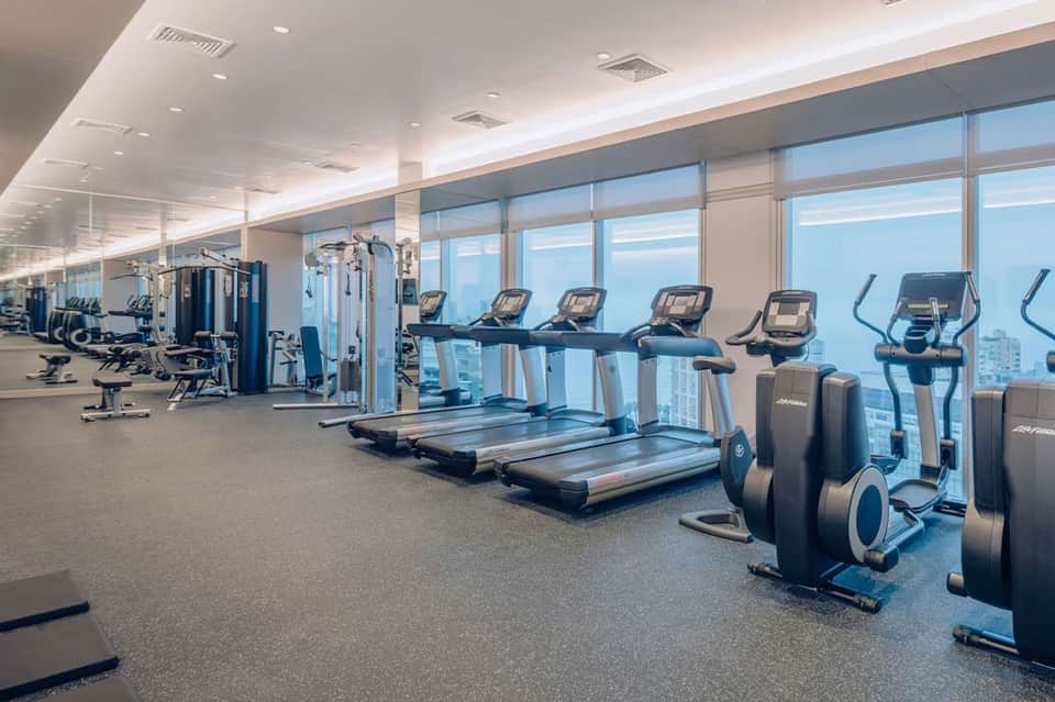 High-rise gym with floor-to-ceiling windows, treadmills, dumbbells, and cityscape views during daytime