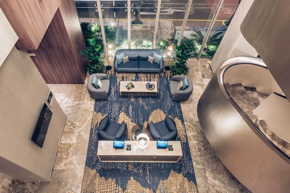 Contemporary hotel lobby with gray seating, large windows, plants, and circular elements