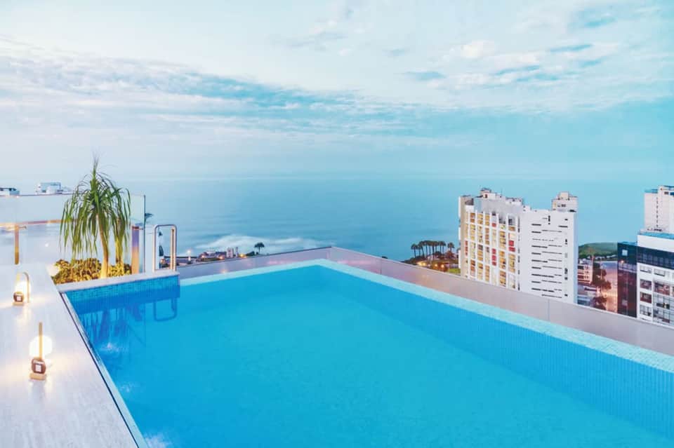 Rooftop infinity pool with turquoise water overlooking ocean, buildings, and coastal horizon at dusk