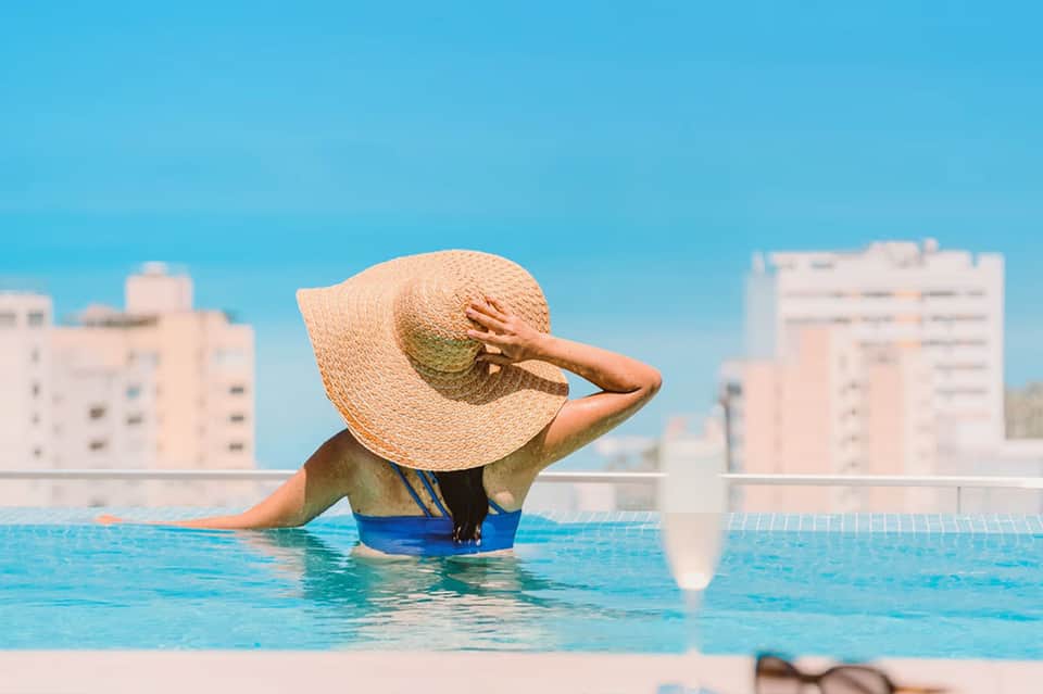 Woman relaxing in rooftop infinity pool wearing straw hat overlooking city buildings and blue sky