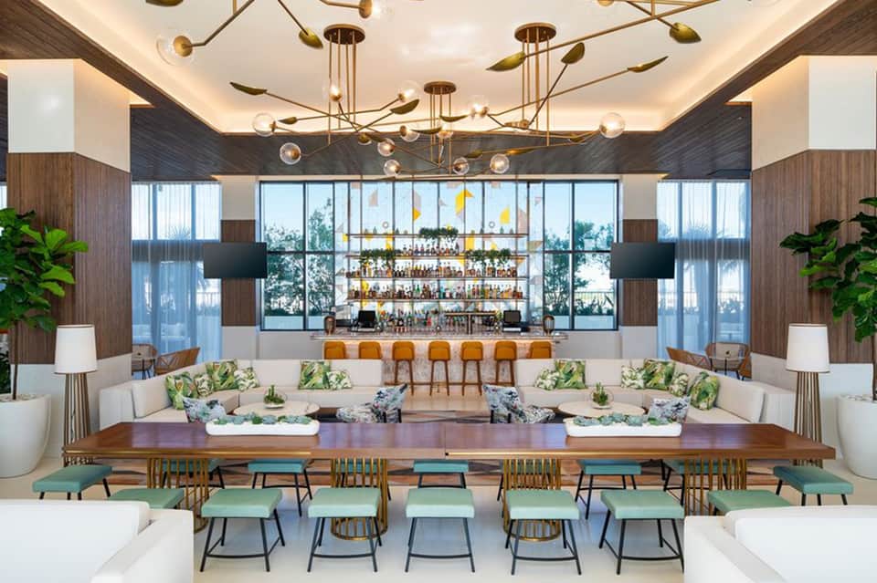 Modern hotel lounge with wooden furnishings, gold pendant lighting, full bar, and tropical plants