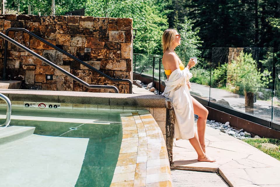 Woman in white robe holding drink by resort hot tub surrounded by stone walls and greenery