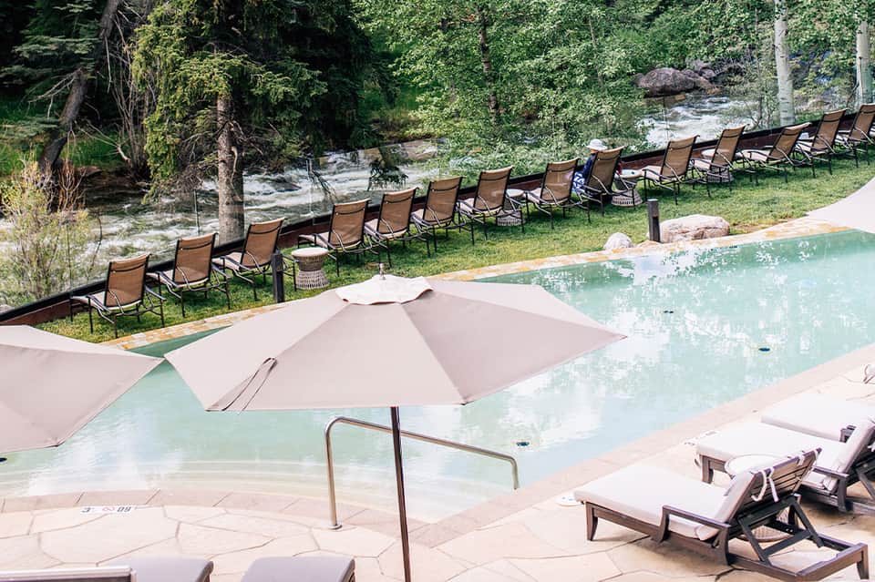 Natural pool surrounded by forest, lounge chairs, umbrellas, and creek with lush greenery