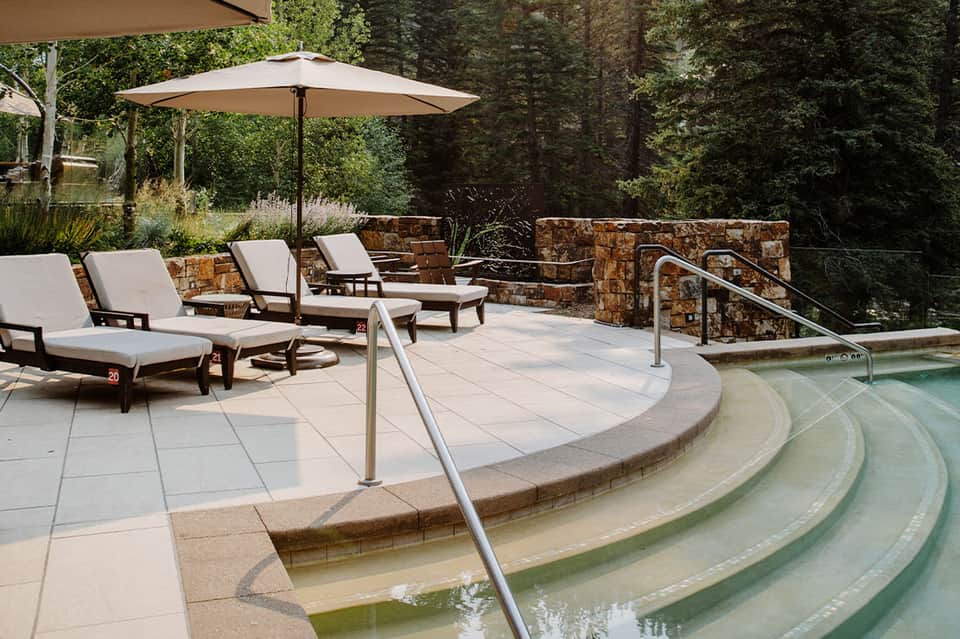 Luxury outdoor hot spring with lounge chairs, umbrella, stone walls, and forest backdrop
