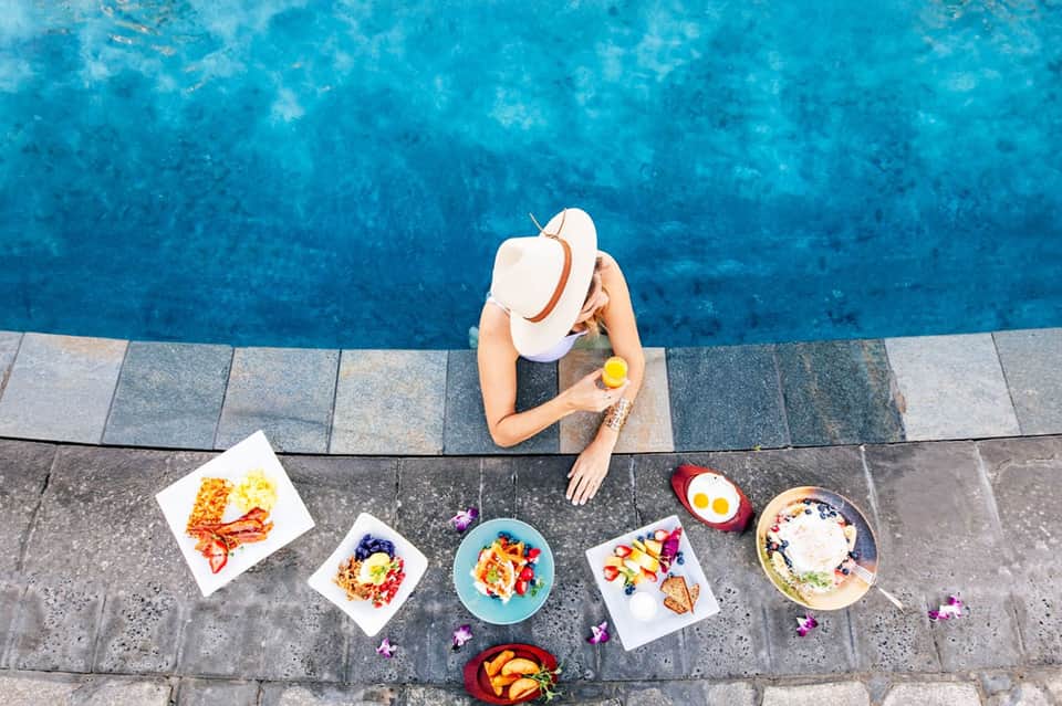 Overhead view of woman at pool edge with breakfast plates and fresh fruit