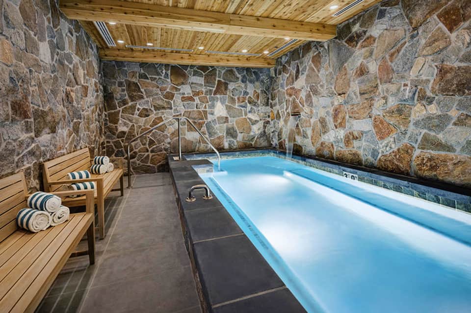 Indoor spa pool with stone walls, wooden ceiling, and lounge chairs with rolled towels
