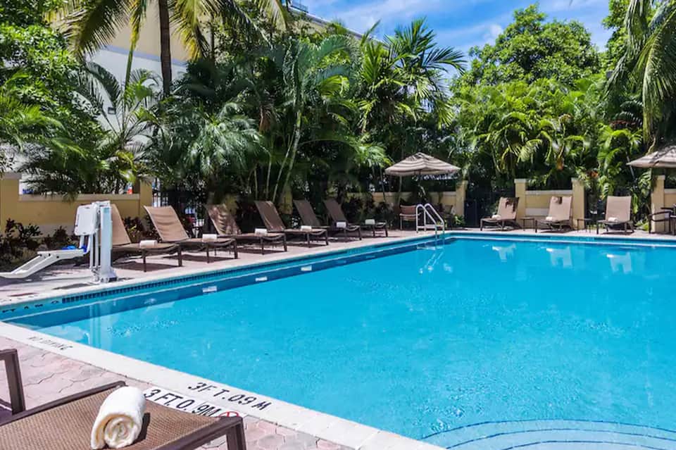 Resort swimming pool with diving board, lounge chairs, tropical palms, and tiki umbrellas