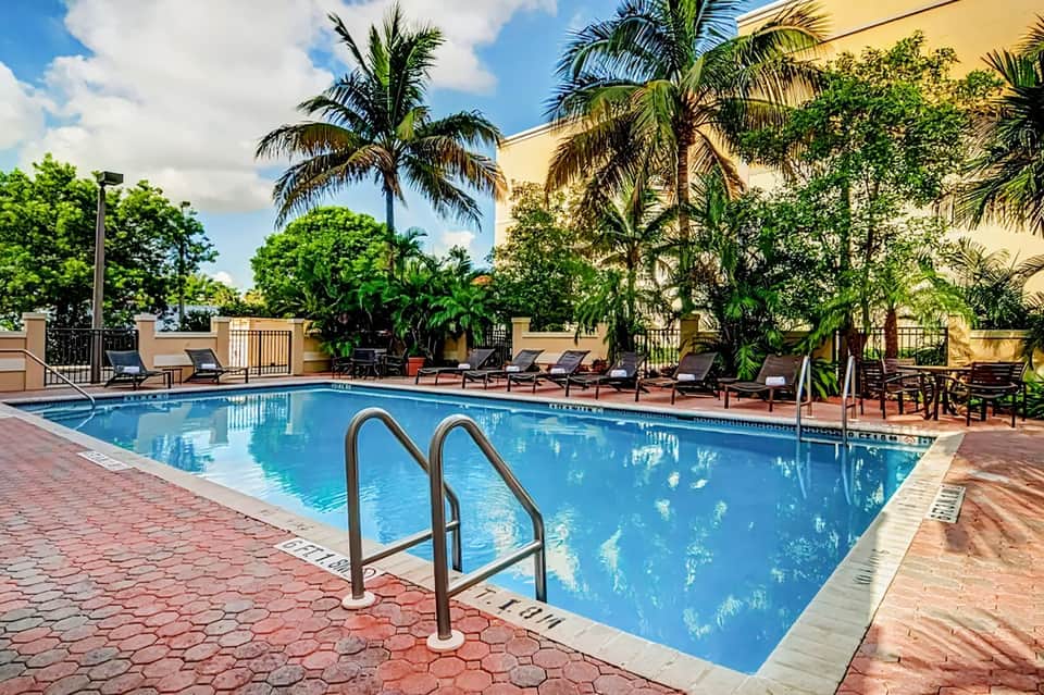 Resort swimming pool with ladder, lounge chairs, palm trees, and tropical courtyard setting