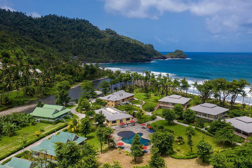 Aerial resort view with multiple bungalows, central pool, lush greenery, and dramatic rocky coastline backdrop