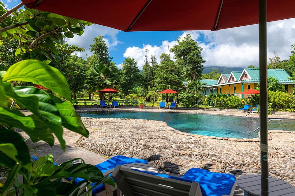Tropical resort beach with colorful cabanas, turquoise water, lush vegetation, and red umbrella shade