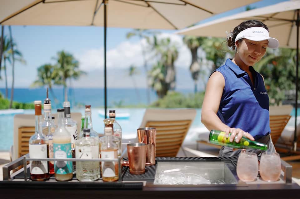 Poolside bar service with bartender preparing drinks under umbrella with ocean and palm trees visible in background