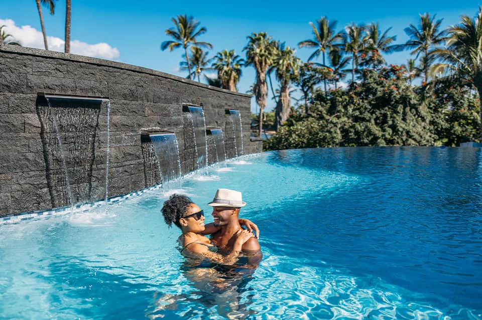 Couple enjoying a modern resort pool with waterfall features, stone walls, and palm trees in background