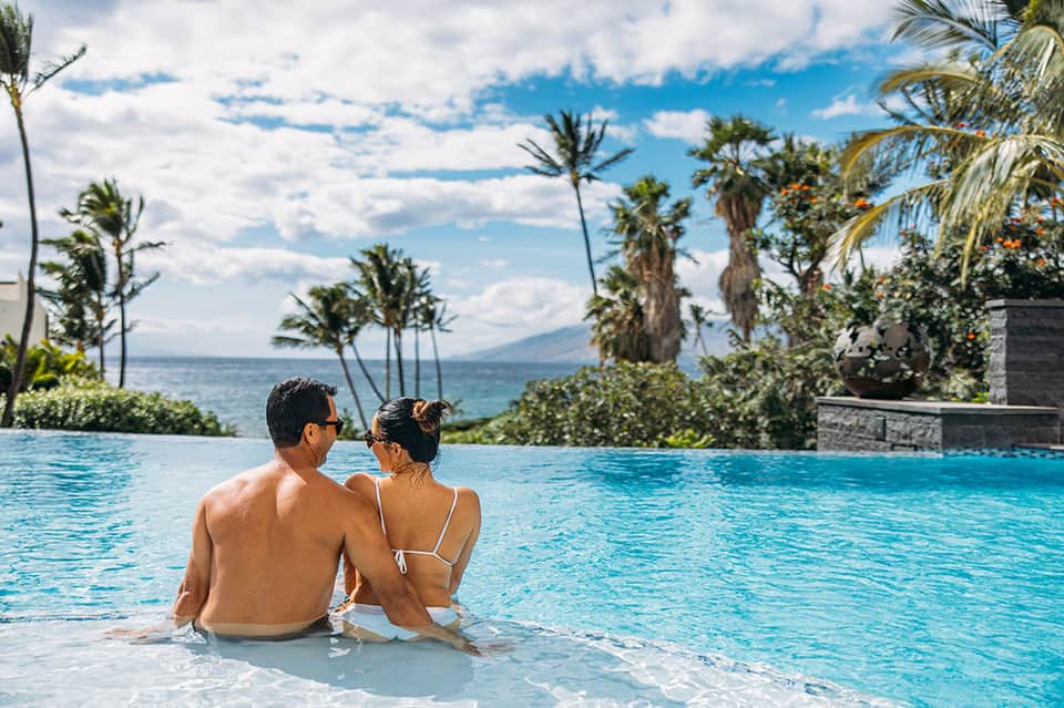 Couple relaxing in infinity pool with ocean view, palm trees, and mountains in background