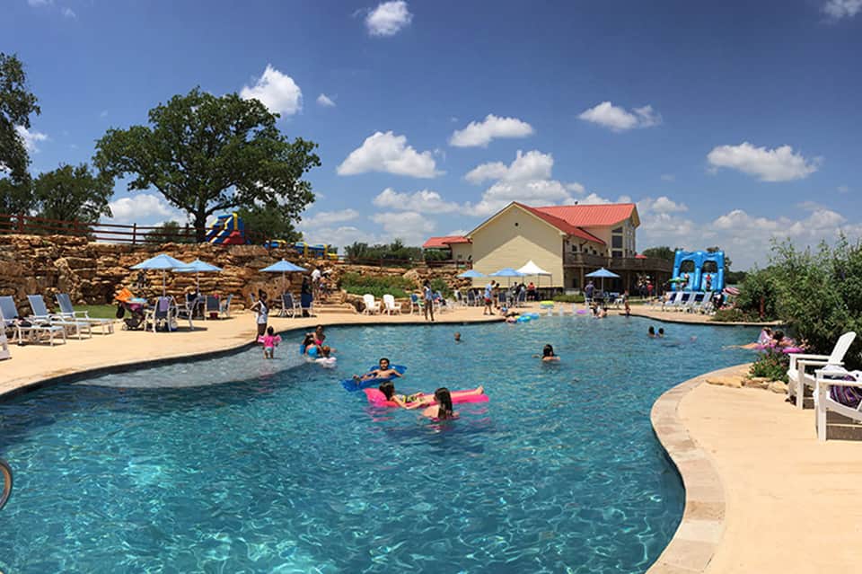 Resort swimming pool with families, sandy area, blue umbrellas, building with red roof in background