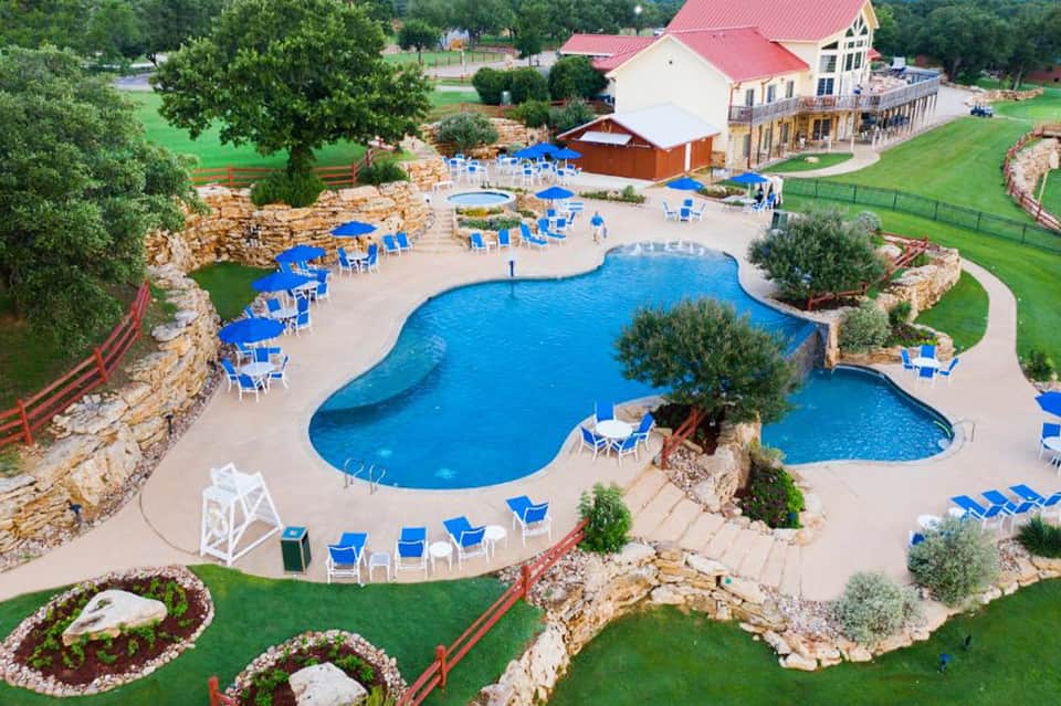 Aerial view of resort pool complex with blue chairs, landscaped grounds, and farmhouse-style main building