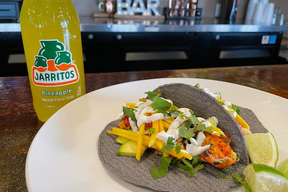 Two charcoal fish tacos with yellow mango strips, lime wedge, and Jarritos pineapple soda on bar counter