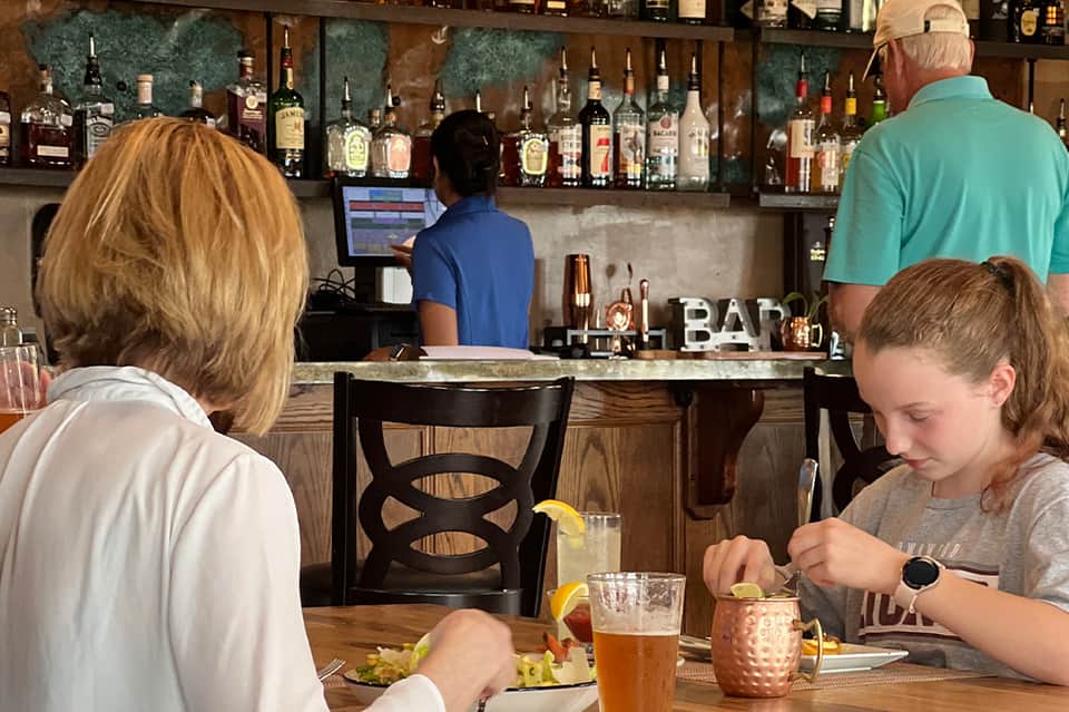 Diners at a bar with cocktails, featuring a well-stocked bar with bottles in background