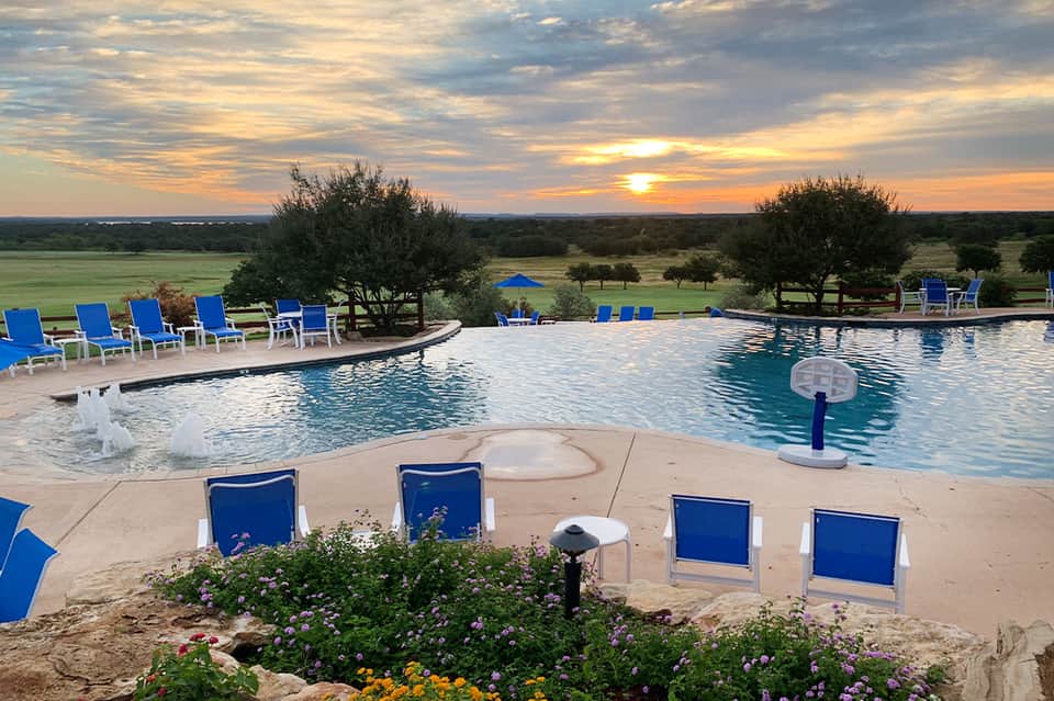 Infinity pool at sunset with blue lounge chairs, flowering gardens, scenic countryside views, and golden sky