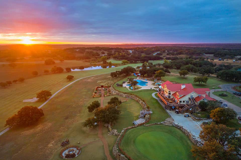 Aerial sunset view of sprawling ranch with red-roof main house, pool, green pastures, and rolling countryside.