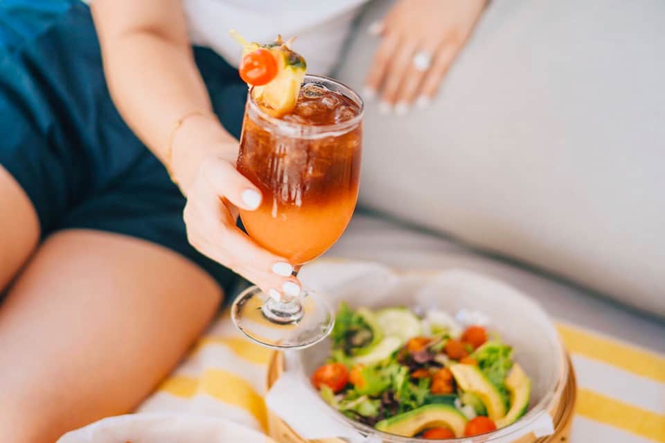 Person holding tropical cocktail with pineapple garnish next to fresh salad on white plate