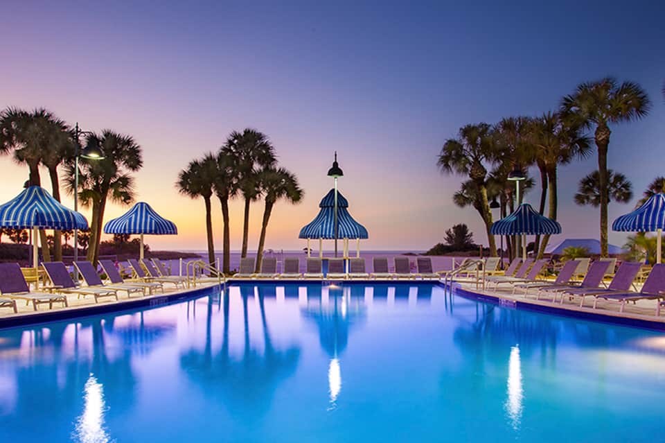 Resort pool at dusk with blue striped umbrellas, palm trees, lounge chairs, and illuminated water