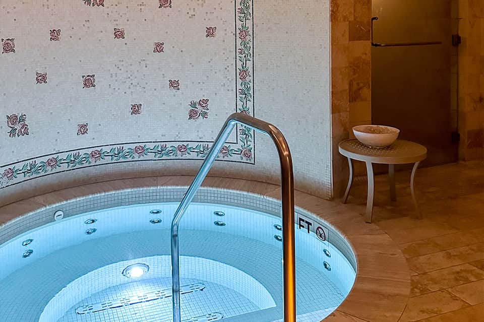 Spa soaking tub with decorative tile work and small wooden table in luxury spa setting