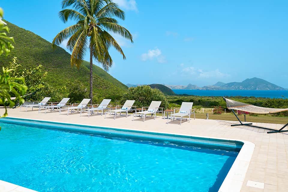 Resort swimming pool with white loungers overlooking lush green hillside and ocean bay view