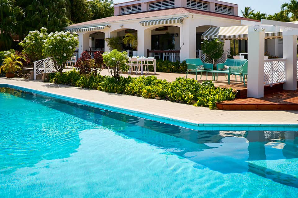Tropical resort pool with turquoise water, white cabana structure, striped awnings, lush gardens, and lounge seating.