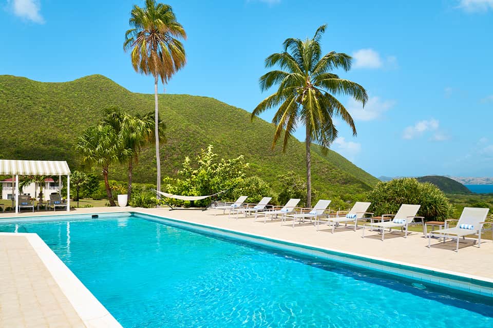 Resort swimming pool with turquoise water, palm trees, and lush green hillside backdrop