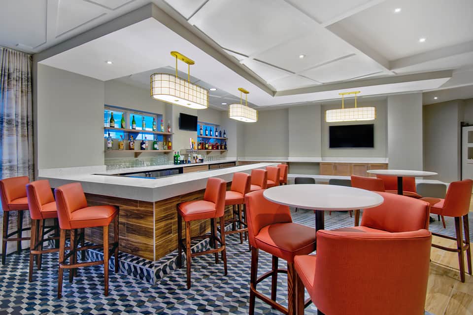 Modern hotel lounge with full bar, orange upholstered seating, gold pendant lights, and white bar counter