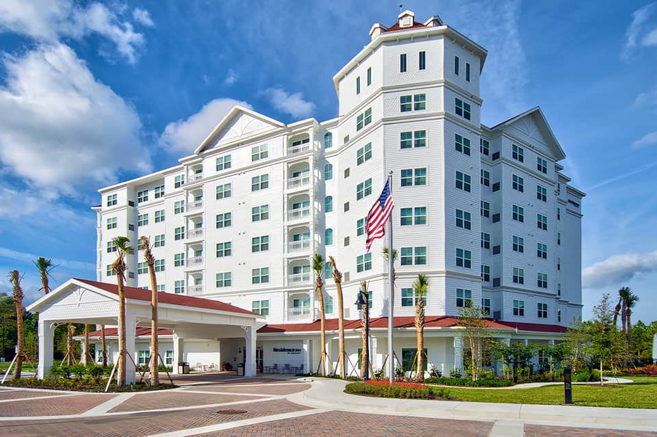 White Victorian-style hotel building with green shutters, central tower, palm trees, and American flag