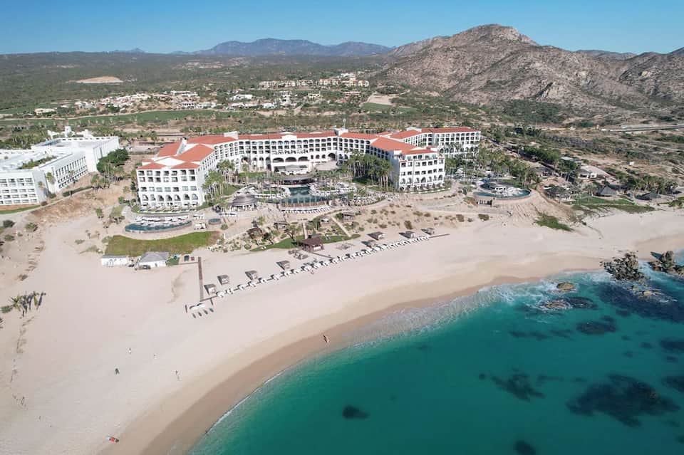 Aerial view of beachfront resort with red roof, multiple pools, and mountain backdrop