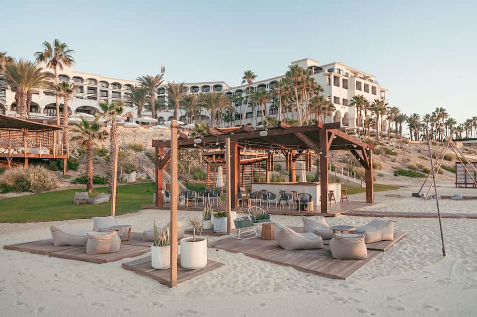 Beachfront resort with palm trees, wooden pergola structures, lounge seating, and hotel buildings