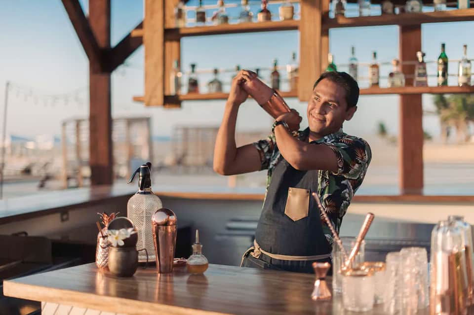 Bartender preparing tropical cocktail behind beachfront bar with ocean view in background