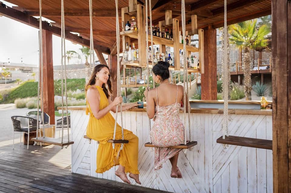 Two women on rope swings in wooden pavilion with ocean view, palm trees, and coastal landscape visible