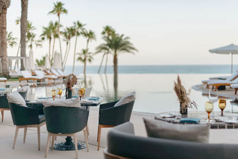 Beachfront dining area with palm trees, ocean view, elegant table settings, and white lounge furniture