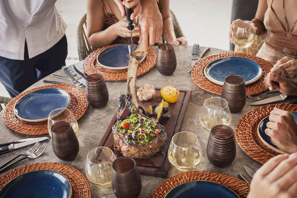 Group dining at outdoor table with woven placemats, blue plates, grilled meat centerpiece, and white wine glasses