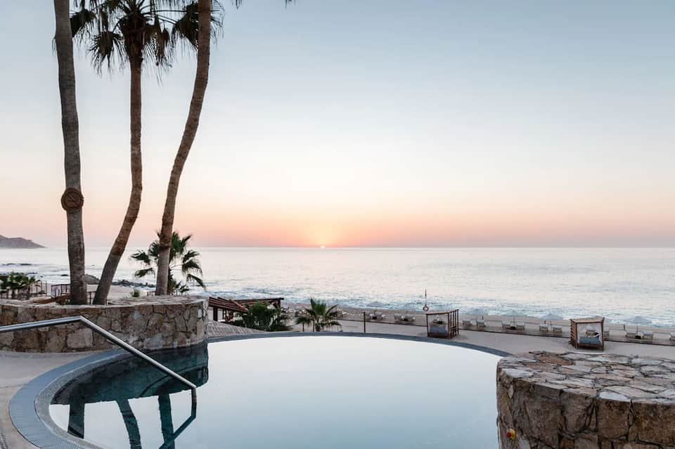 Peaceful beachfront pool at sunset with palm trees, ocean view, and beach loungers visible below
