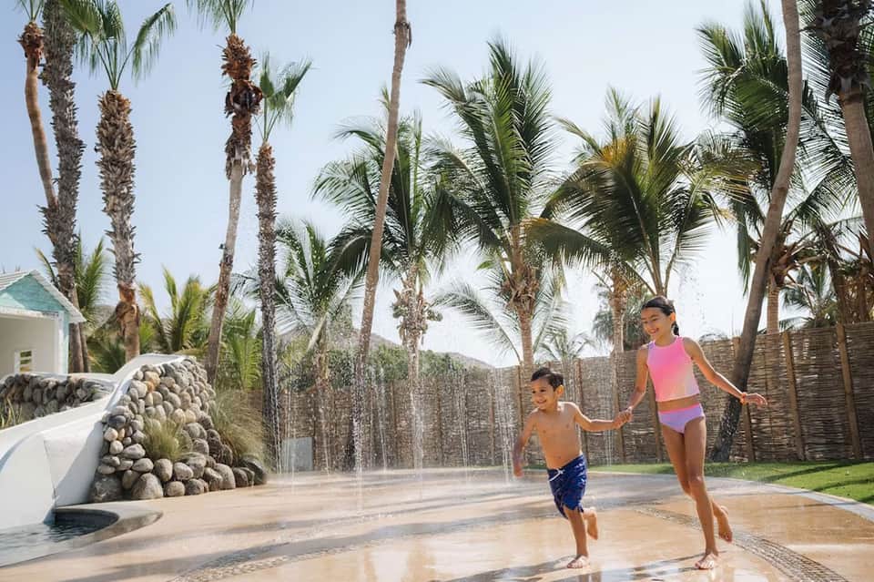 Children playing in shallow splash pad surrounded by palm trees and resort buildings