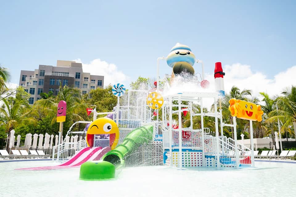 Colorful water splash pad with cartoon character sculptures and white playground structure surrounded by palm trees