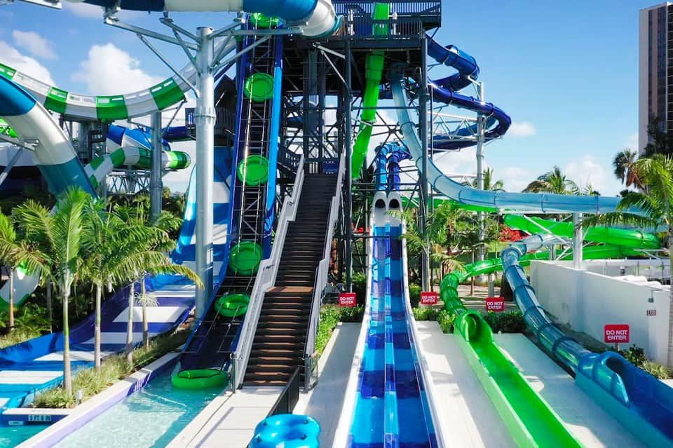 Colorful water slides with blue and green tubes at an outdoor waterpark surrounded by palm trees