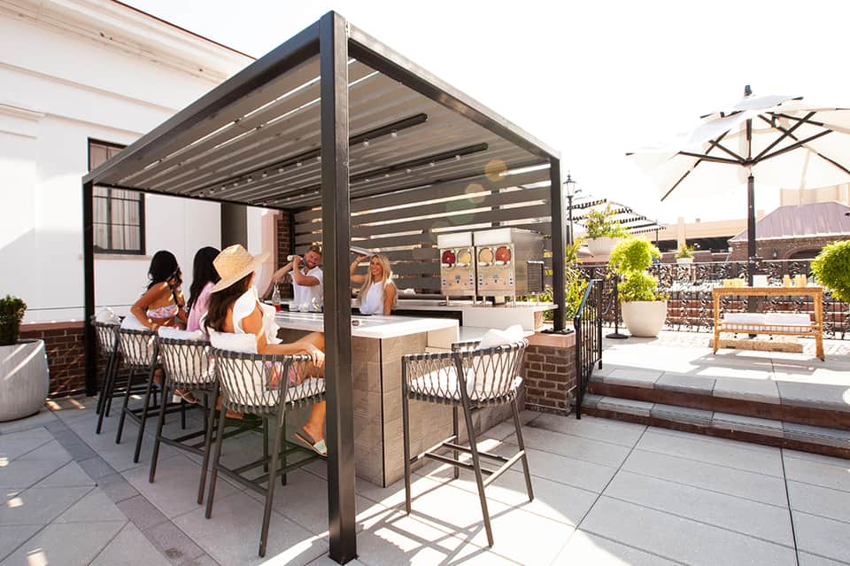Rooftop bar with pergola structure, patrons at counter, city skyline views, and umbrella seating