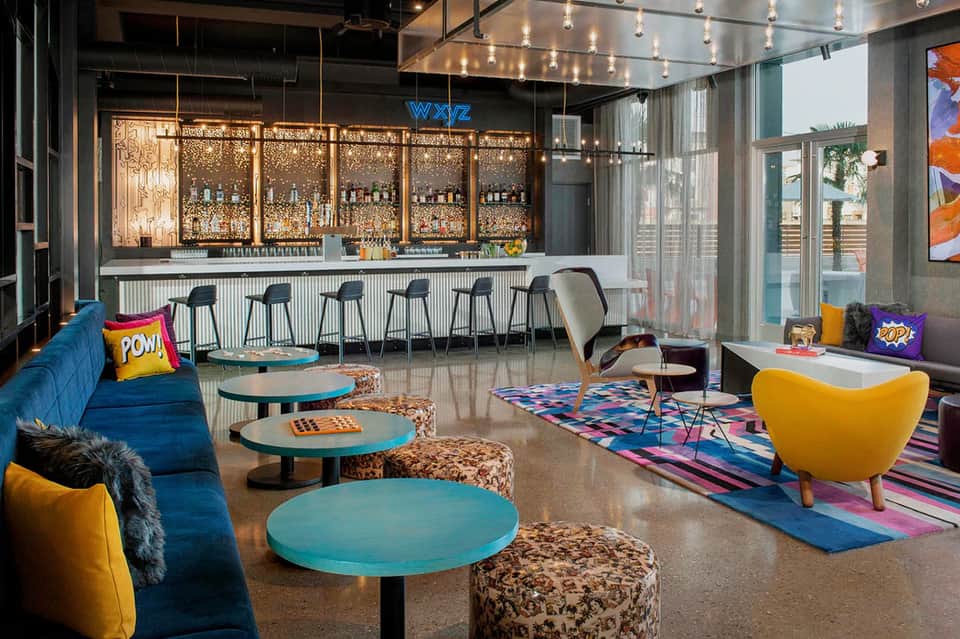 Contemporary hotel lobby with blue seating, colorful accents, glass bar, and pop art decor