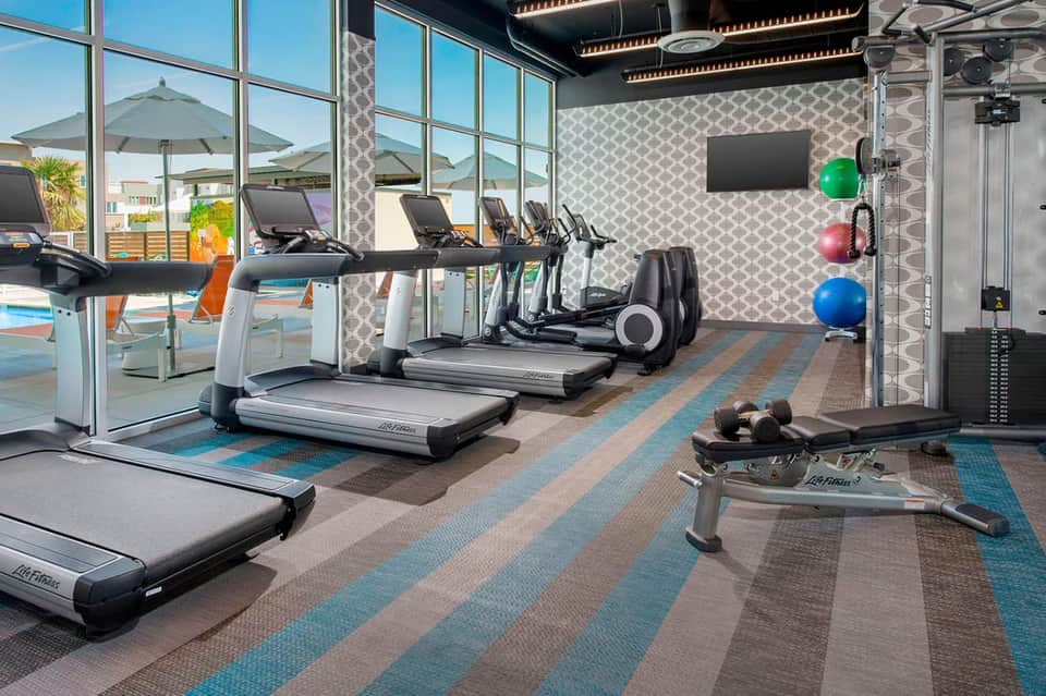 Fitness center with treadmills, stationary bikes, exercise equipment, and large windows overlooking the pool area.