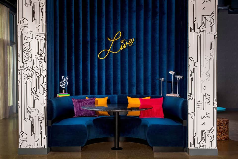 Modern lounge with bold blue velvet curved seating, neon yellow 'Live' sign, and colorful pillows