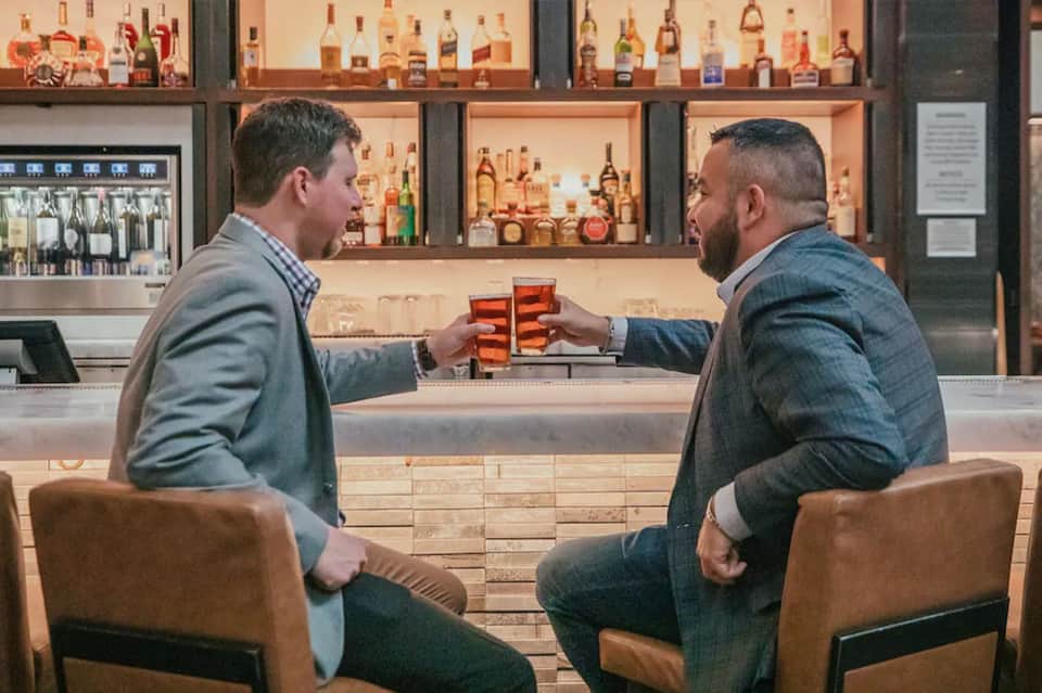 Two men toasting cocktails at upscale bar with extensive liquor collection and wooden shelving