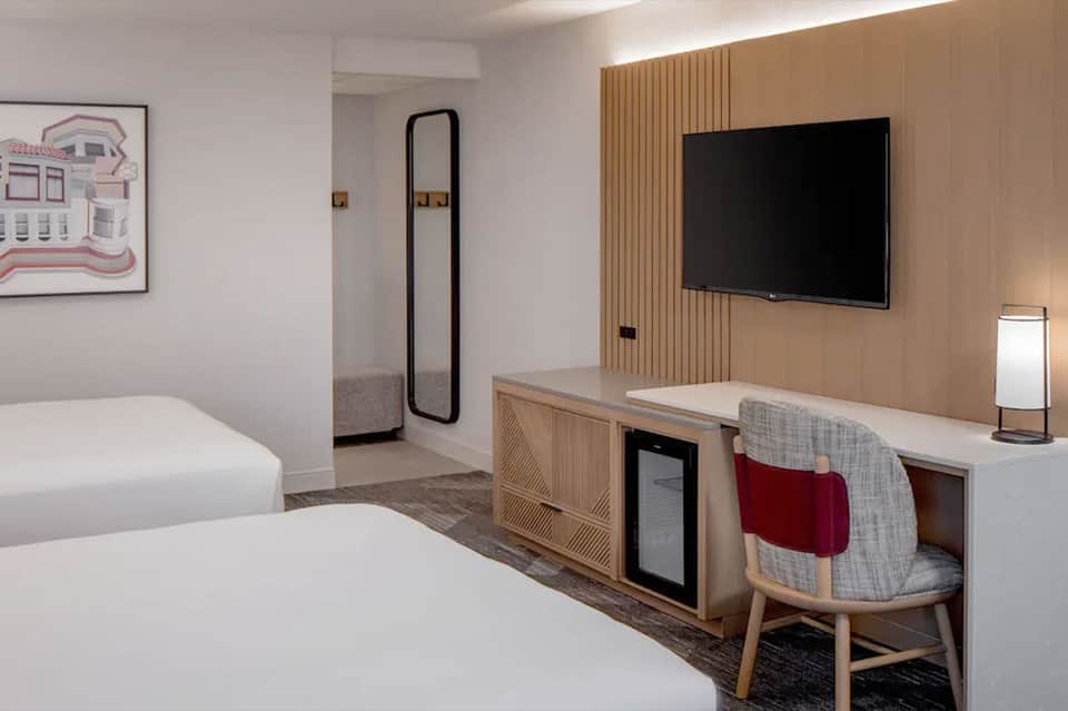 Modern hotel room with two beds, desk area, wall-mounted TV, and natural wood accents