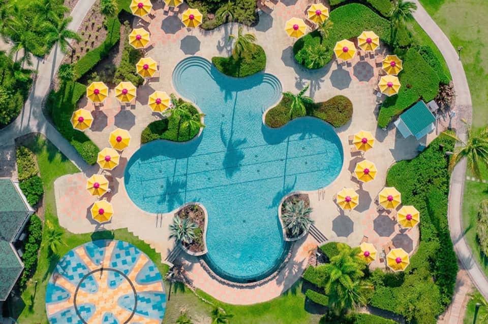 Aerial view of resort pool complex with yellow umbrellas, surrounding landscape, and curved pathways