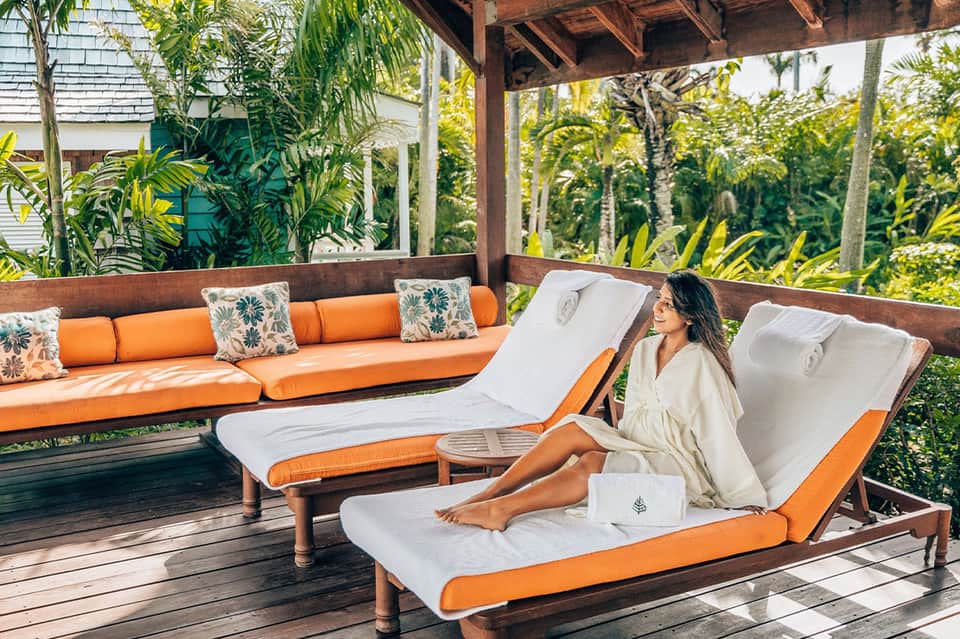 Woman relaxing on orange and white chaise lounge under wooden pergola surrounded by tropical palm plants